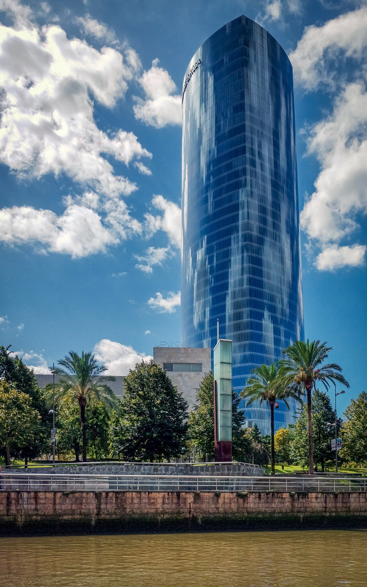 The glass shaft of the Iberdrola Tower against a sky of broken cloud, palms at its foot