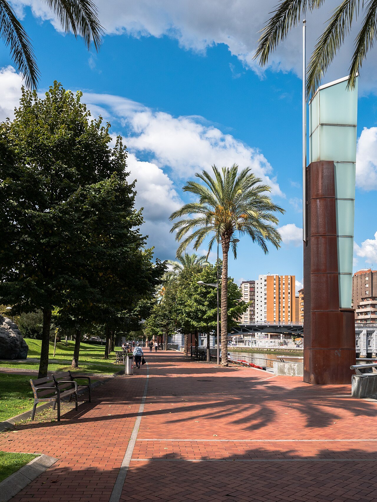 A palm-lined walk on the riverside promenade, a rusted steel sculpture beside the path