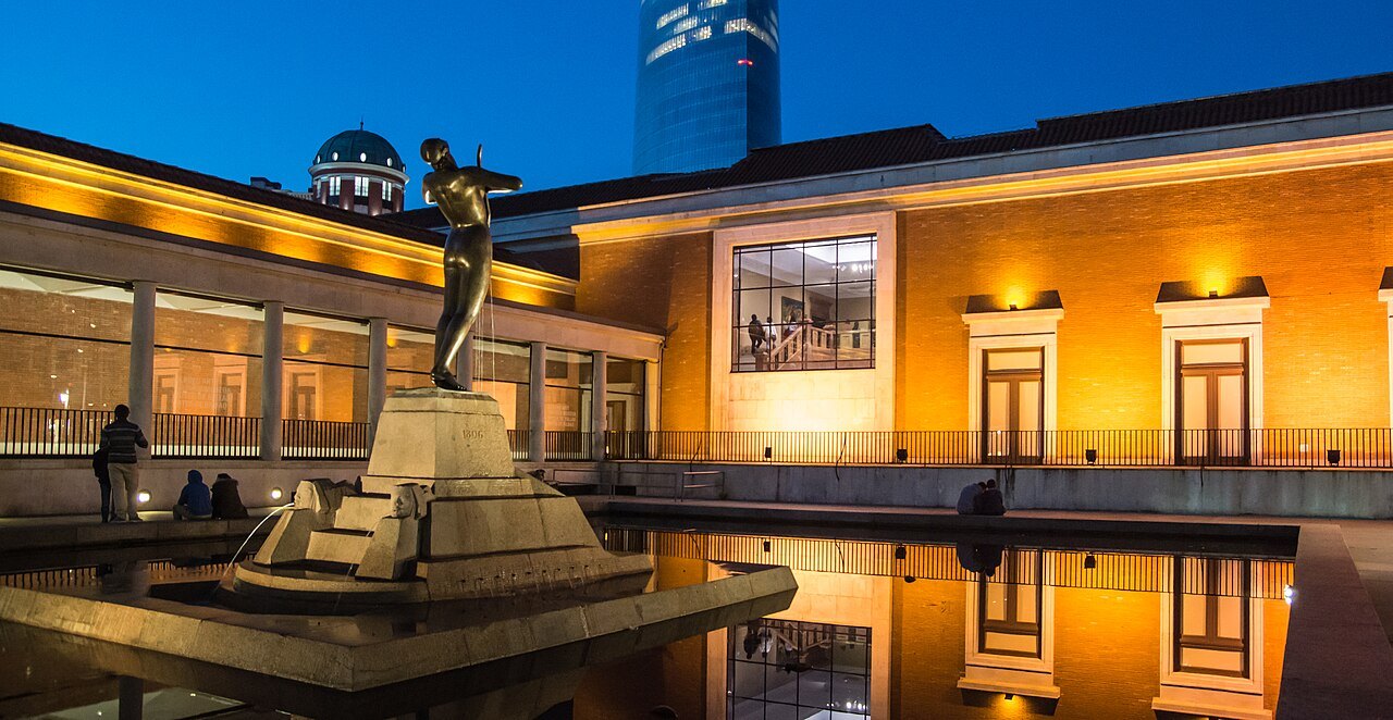 The Fine Arts Museum lit at dusk, a bronze figure reflected in the pool in front