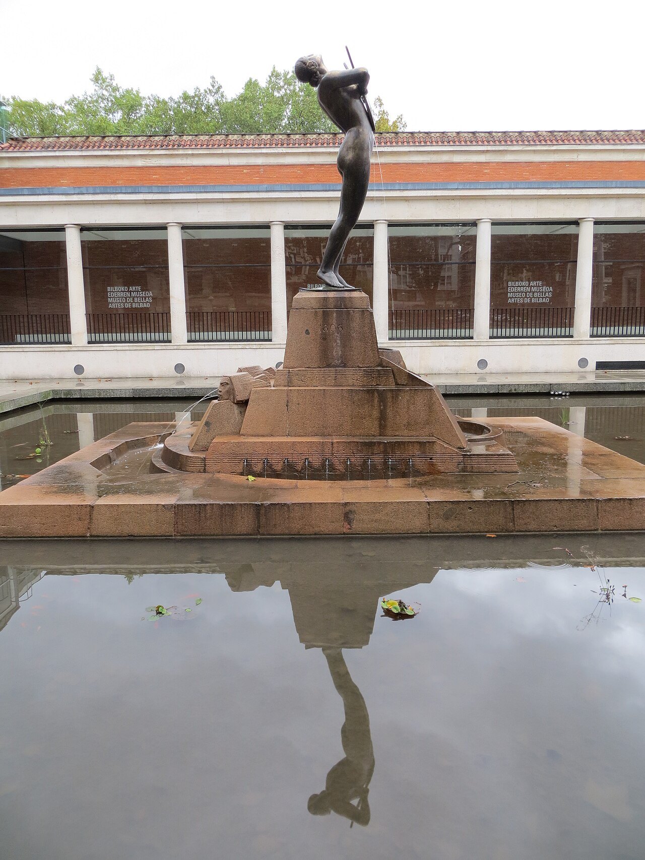 A bronze dancing figure on a stone pedestal reflected in the museum's pool