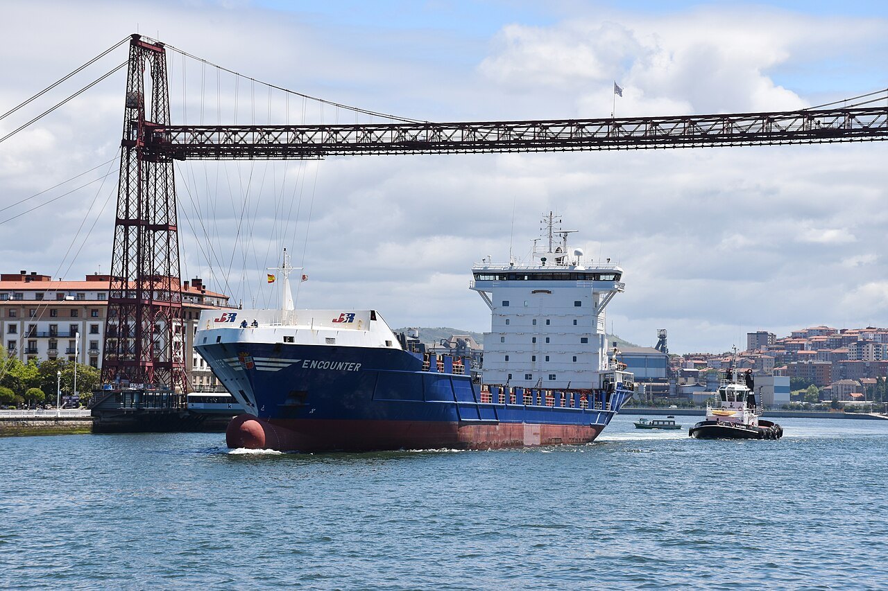 A cargo ship and a tug passing under the Bizkaia Bridge