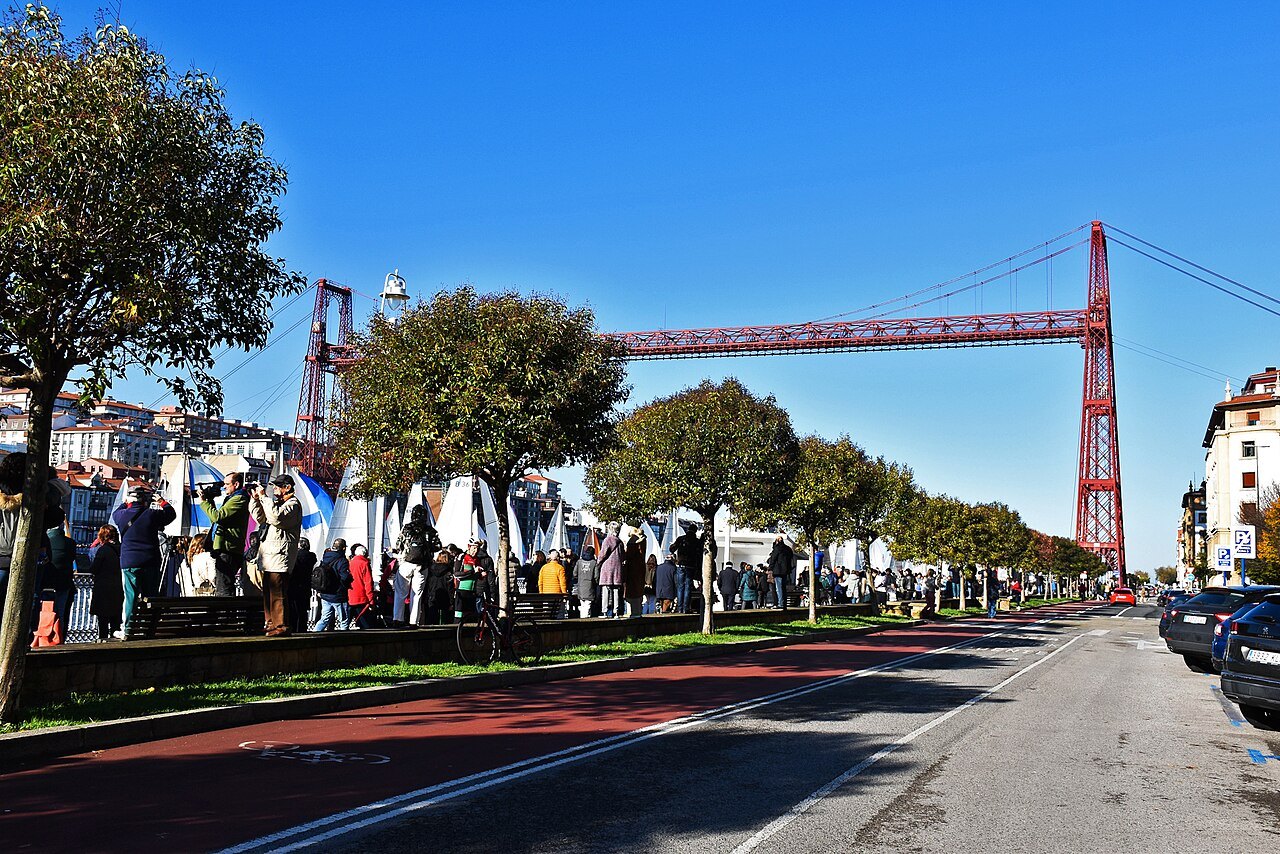 A crowd and market stalls on the tree-lined avenue below the Bizkaia Bridge