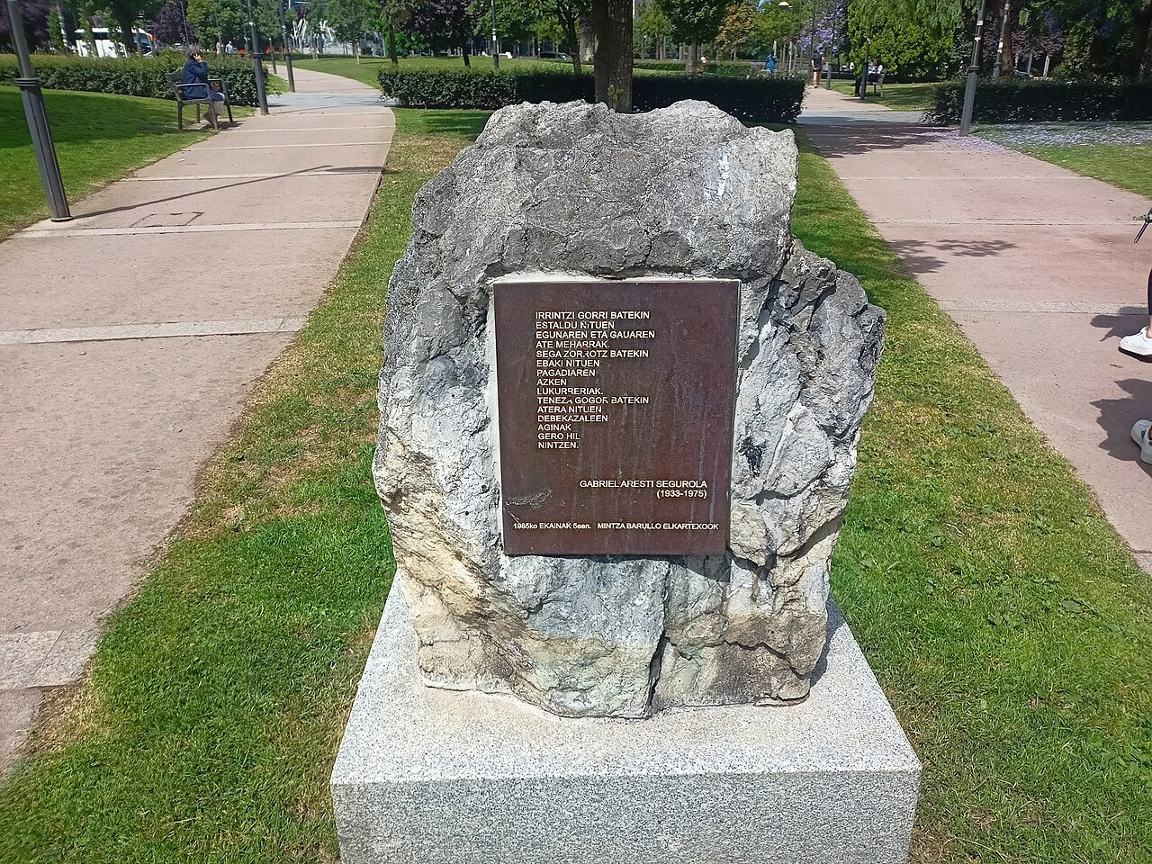 A rough stone carrying a plaque to the poet Gabriel Aresti, beside a park path