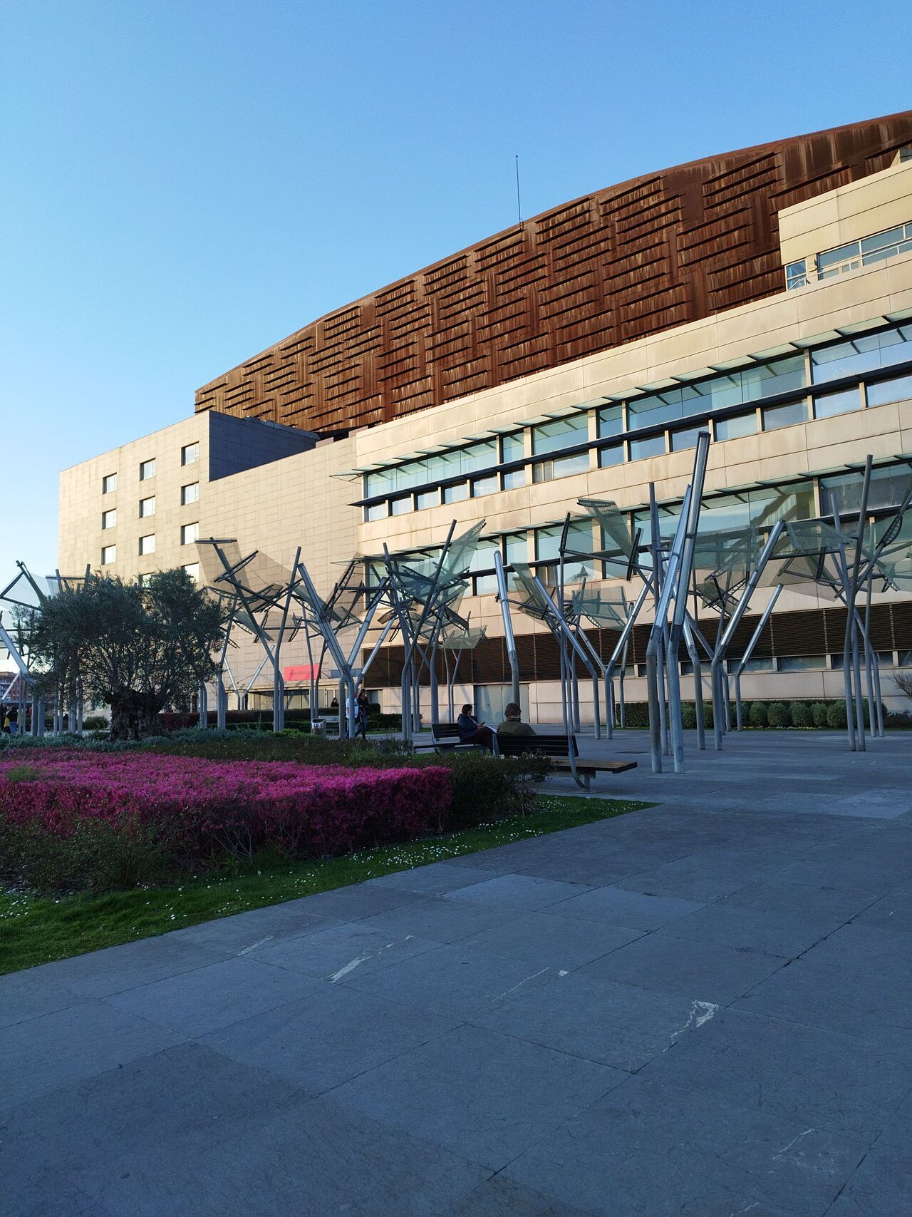 The rusted, curving roofline of the Euskalduna over flower beds and lamp trees