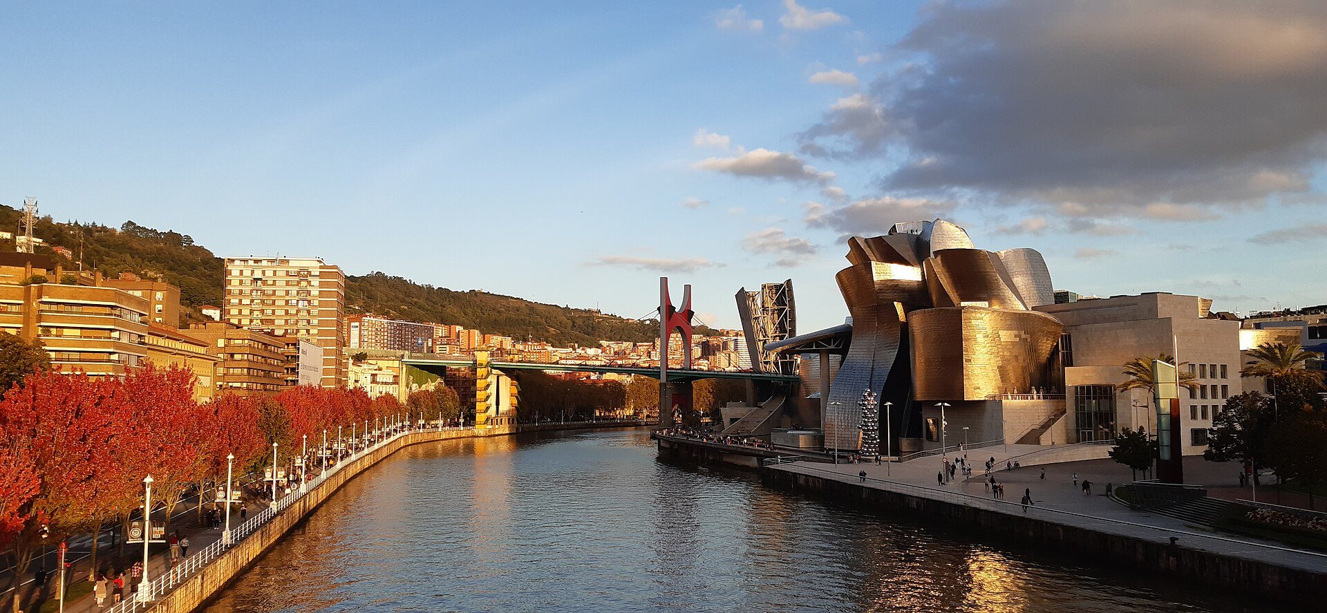 The Guggenheim's titanium in evening sun across the Ría, with the red arch of La Salve Bridge and autumn trees along the bank